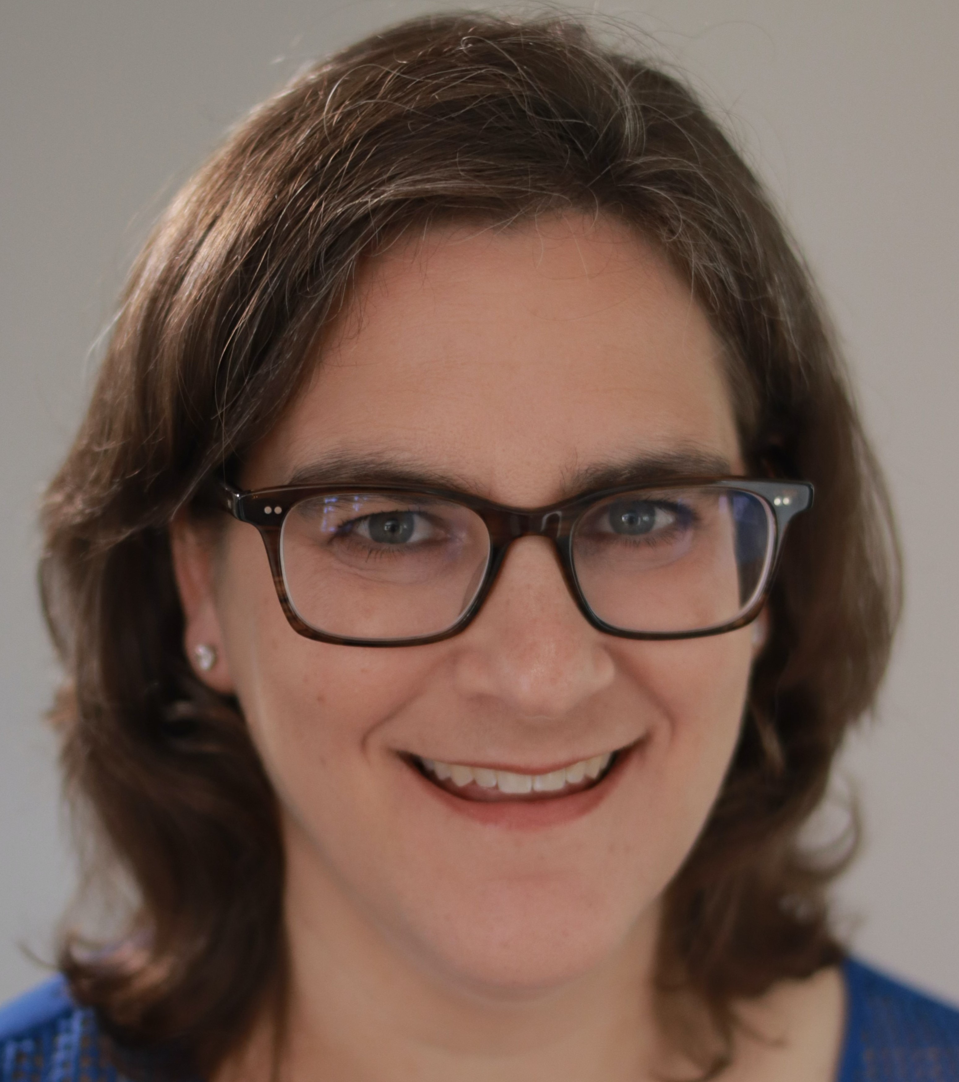 A head shot of Hilary Seligman. Hilary is fair skinned woman with brown hair, and light colored eyes. She is wearing a blue shirt and black glasses, and standing in front of a grey background.