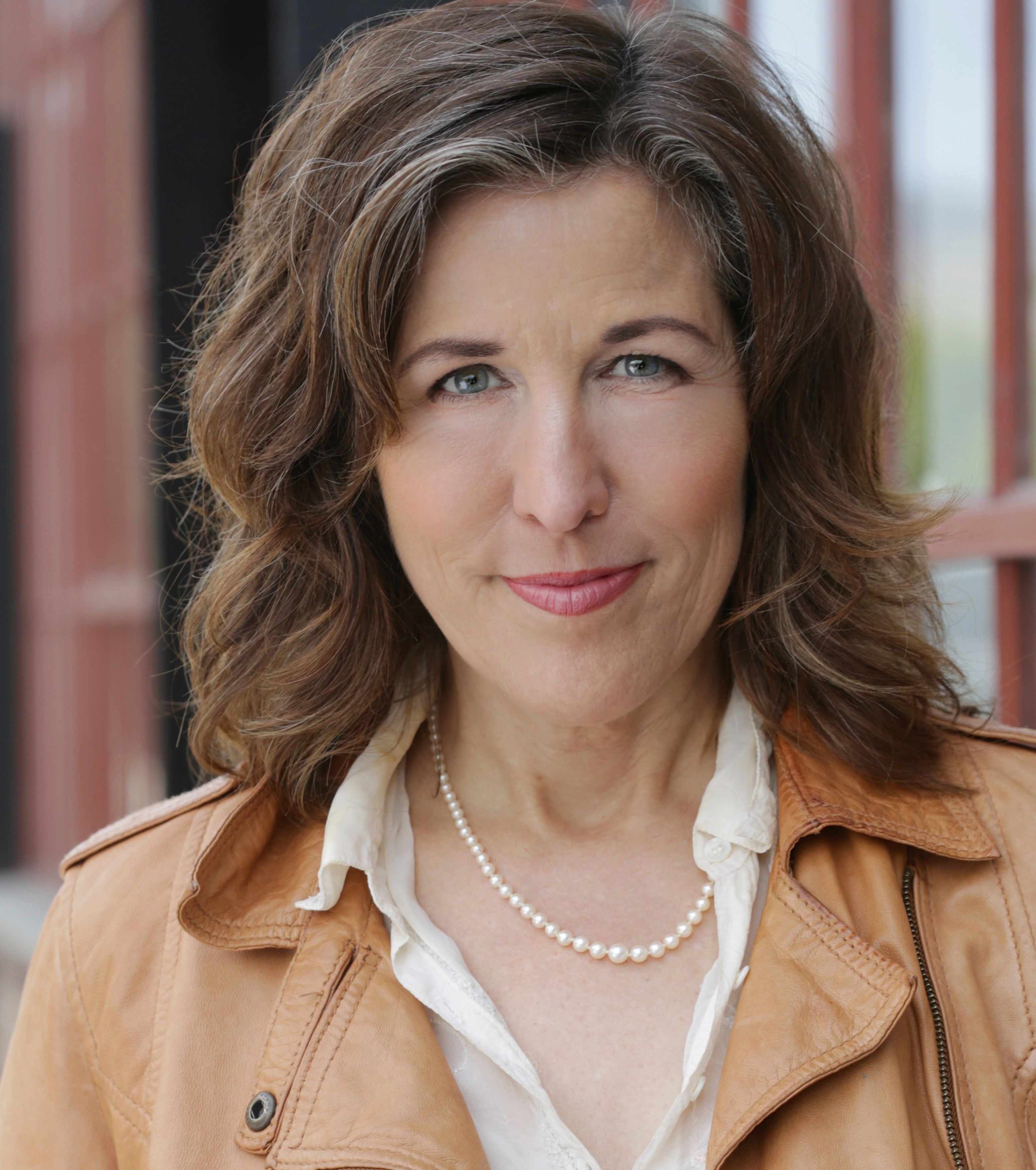 A head shot of Laura Schmidt. Laura is a fair skinned woman with brown hair and blue eyes. She is standing in front of windows on a building. She is wearing a white button up shirt with a tan jacket over it. She is also wearing a pearl necklace.